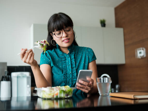 Young businesswoman eating salad in lunch room and using phone stock photo