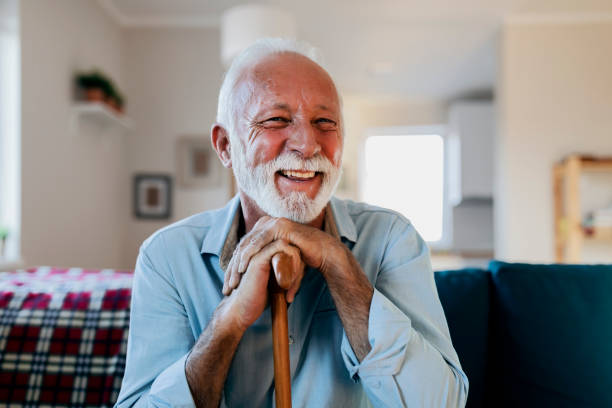 portrait of a happy senior man sitting and holding his walking stick in a nursing home during the morning Portrait of happy senior man sitting at home with walking stock and smiling.Portrait of nice cheerful positive cheery stylish old man wearing checked shirt leaning on cane in white light modern interior studio room new house persons with disabilities photos stock pictures, royalty-free photos & images