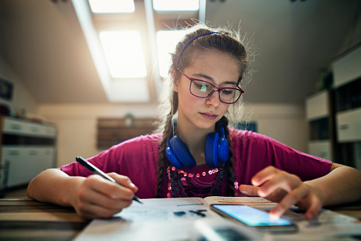 Teenage girl doing homework at home. The girl is using her smartphone and wearing headphone around her neck.
Nikon D850