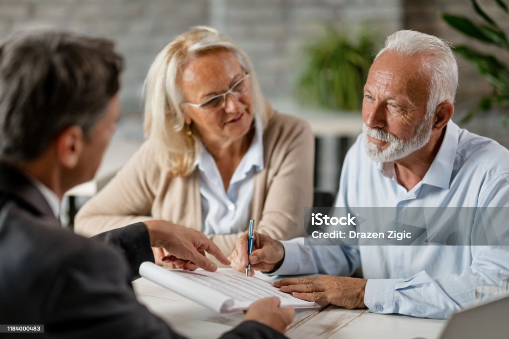 Senior couple signing a contract while having a meeting with insurance agent in the office. Mature couple having a meeting with bank manager and signing lease agreement in the office. Focus is mature man. Will - Legal Document Stock Photo