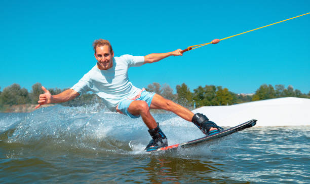 ritratto: allegri wakeboarder dà il segno shaka mentre accelera oltre la fotocamera. - wakeboarding surfing men vacations foto e immagini stock