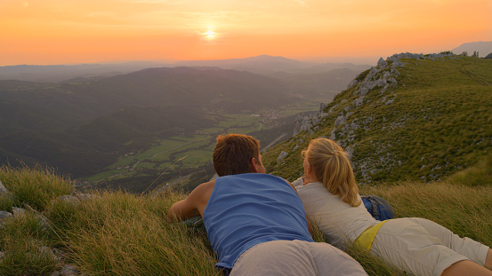 CLOSE UP: Cheerful man and woman on active date watch the beautiful summer sunset illuminate green mountains and valley. Carefree couple enjoying a romantic summer evening in the peaceful mountains.