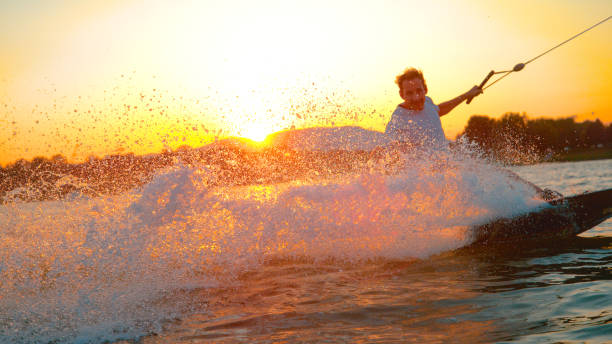 lens flare: il wakeboard allegro spruzza l'acqua del lago alla telecamera al tramonto - wakeboarding surfing men vacations foto e immagini stock