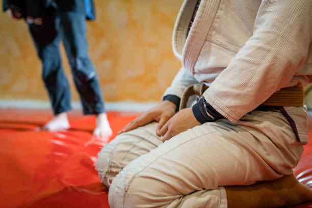 nahaufnahme auf dem mittelteil des bjj brasilianischen jiu jitsu athleten sitzt auf den matten tatami bei der trainingsklasse mit händen auf seinen krawatten tragen weiß gi kimono - ju jitsu stock-fotos und bilder