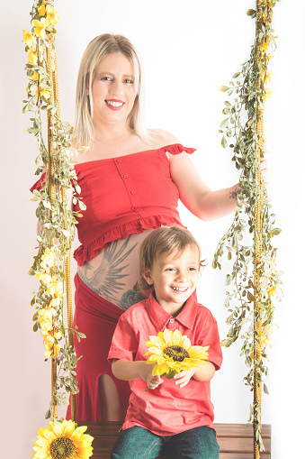 pregnant woman portrait in studio sitting on flowered swing with sunflower and your little son