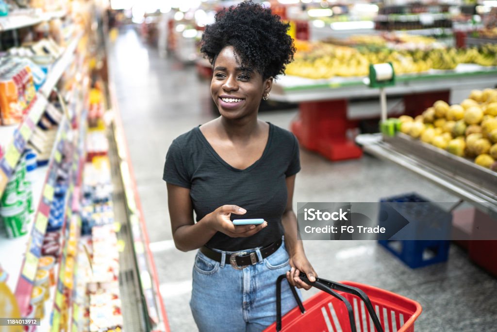 Giovane donna che usa il cellulare e sceglie il prodotto al supermercato - Foto stock royalty-free di Supermercato