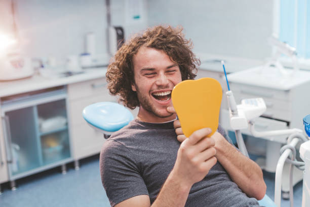 치과 진료소에서 주황색 거울을 들고 곱슬 머리를 한 큰 남자가 치과 의자에 앉아있는 그의 치아를 검사합니다. - dentist office dentists chair dental equipment medical equipment 뉴스 사진 이미지
