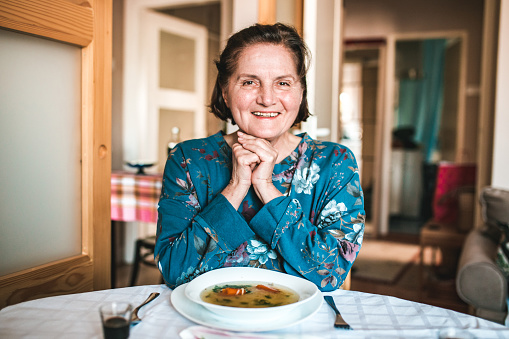 Real Slavic woman is siting at the kitchen table. She is looking at the camera and soup is on the table