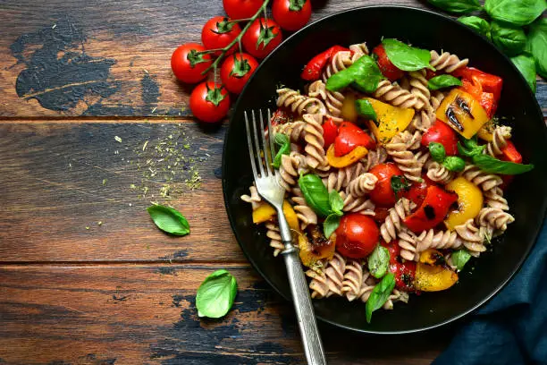 Whole wheat fusilli pasta with grilled vegetables in a black bowl on a wooden background. Top view with copy space.