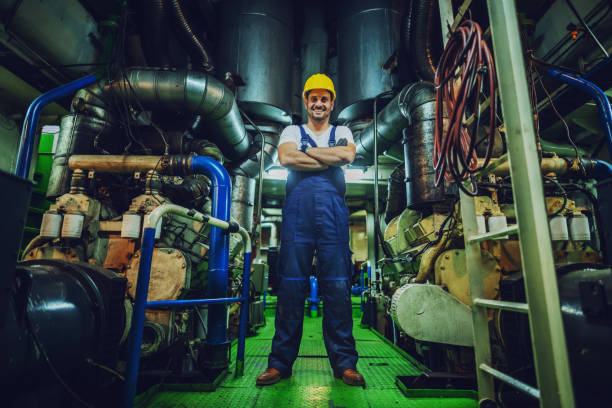 Low angle view of handsome caucasian worker in overall and helmet on head standing in ship next to engine with arms crossed. Low angle view of handsome caucasian worker in overall and helmet on head standing in ship next to engine with arms crossed. motorboat maintenance stock pictures, royalty-free photos & images