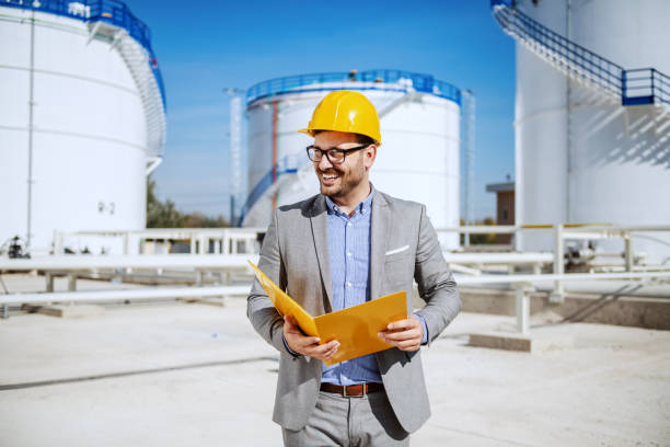 istock Handsome caucasian businessman in suit standing outdoors and holding folder. In background are oil storage tanks. 1184040739
