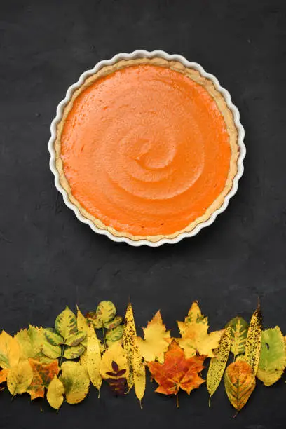 Bright orange pumpkin pie and dry leaves looking like sunset over autumn forest on black background. Healthy natural sweets concept overhead view