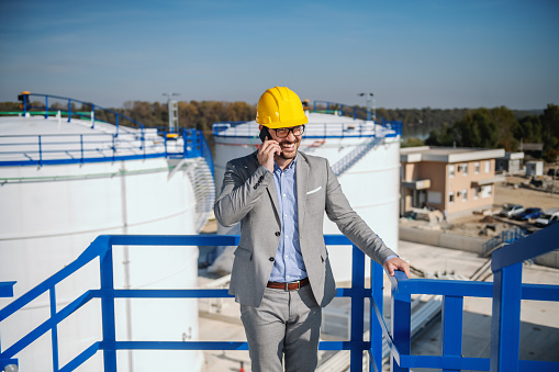 Handsome caucasian businessman in suit and with helmet on head talking on the phone while standing outdoors. In background are oil tank storage.