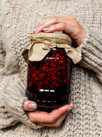 Glass jar with raspberry jam in the hands of girls. A girl in a wool sweater holding a jar of jam. Folk medicine.