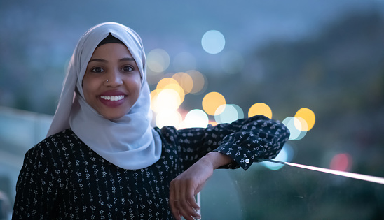 African  Muslim woman in night  at balcony  smiling at camera with city bokeh lights in background