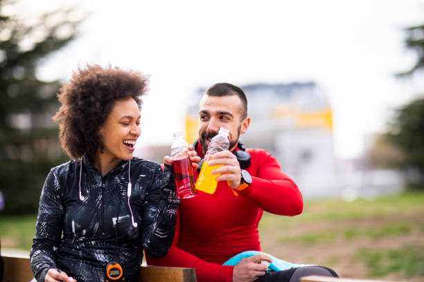 istock Young athletic diverse couple sitting next to each other on a bench in a park and doing a toast with their energy drinks 1184137032