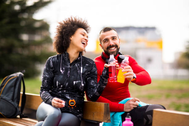 Two young athletes in sports clothing putting their energy drink bottles together while sitting on a bench in a public park Young athletic couple sitting next to each other on a bench in a park and doing a celebratory toast with their energy drinks. sport drink stock pictures, royalty-free photos & images