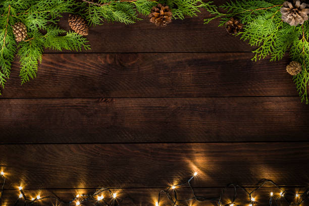 Christmas decoration with copy space on a rustic wooden table Top view of a dark brown wooden table with some pine twigs and pine cones at the top of the image and a Christmas light string at the botton. Objects are disposed on a frame shape leaving a useful copy space at the center of the image. 
Low key DSLR photo taken with Canon EOS 6D Mark II and Canon EF 24-105 mm f/4L christmas pine cone frame branch stock pictures, royalty-free photos & images