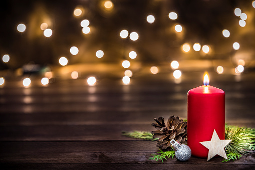 Front view of a red christmas candle surrounded by a silver christmas ball, a pine cone, a wooden star shape and pine twigs on a rustic wooden table. At the background are some defocused christmas lights. The objects are at the right side of the image leaving a useful copy space at the opposite side. \nLow key DSLR photo taken with Canon EOS 6D Mark II and Canon EF 24-105 mm f/4L