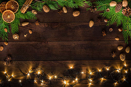 Top view of some pine twigs, Christmas lights, star anise, almonds and nuts, dried oranges, cinnamon sticks, pine cones on a rustic wooden table. The Christmas lights are at the botton of the image and the other objects are disposed on a frame shape leaving a useful copy space at the center of the image.\nLow key DSLR photo taken with Canon EOS 6D Mark II and Canon EF 24-105 mm f/4L
