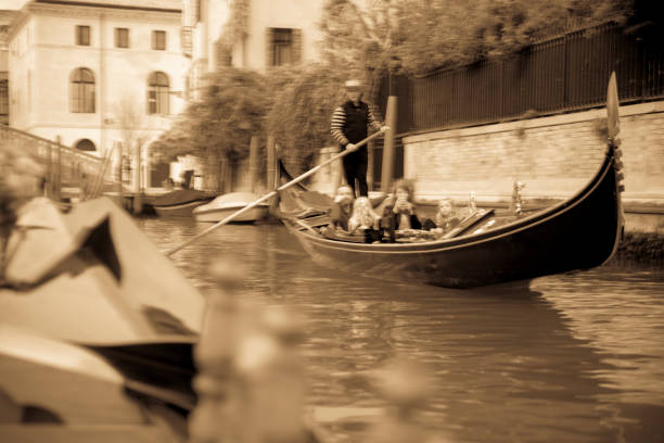 Venice Gondola canal trip with POV from the gondola of bridges and other gondolas near the Bridge 'Ponte Dei Greci'. The gondoliers are in traditional dress and steering the boat past moored boats and other gondolas and water taxis. It is nearly dark.