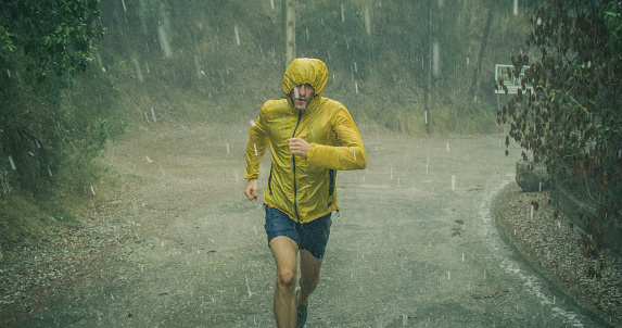 Athletic muscular young man in sports outfit is jogging in the street on a rain