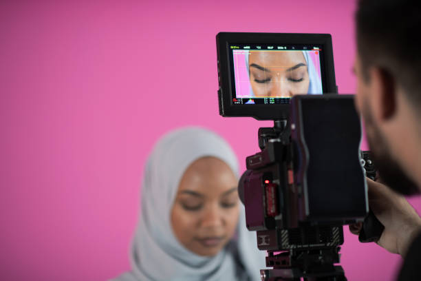 videographer in pink studio recording video on professional camera by shooting female muslim woman male videographer in digital studio recording video on professional camera by shooting female Muslim woman wearing hijab scarf plastic pink background muslim photographer stock pictures, royalty-free photos & images