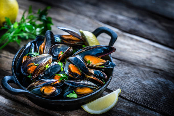 High angle view of steamed mussels in a cast iron cooking pan shot on rustic wooden table. The composition is at the left of an horizontal frame leaving useful copy space for text and/or logo at the right. Selective focus on the mussels. Predominant colors are black, orange and brown. XXXL 42Mp studio photo taken with Sony A7rii and Sony FE 90mm f2.8 macro G OSS lens