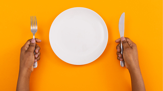 Dinner place setting. White empty plate and cutlery in black woman's hands on orange background, top view