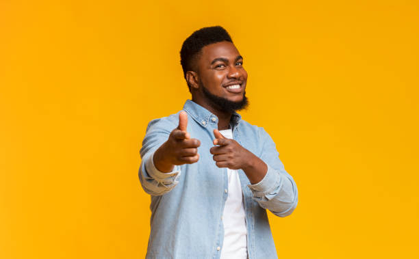 Cheerful bearded black guy pointing fingers at camera Smiling bearded african american man pointing fingers at camera over yellow background with copy space charming stock pictures, royalty-free photos & images