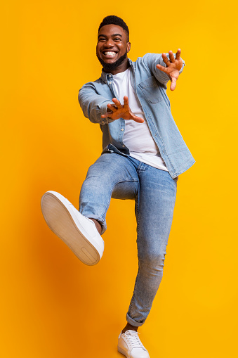 Funny african american guy dancing and extending his hands to camera over yellow studio background, low-angle view
