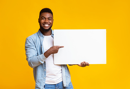 Handsome african american man pointing at blank white advertising board in his hands, yellow studio background with free space