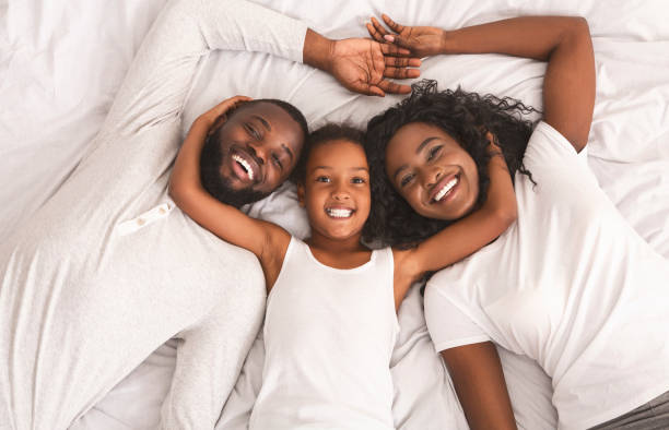 улыбаясь черная мать, отец и дочь, лежащие на кровати вместе - couple affectionate relaxation high angle view стоковые фото и изображения