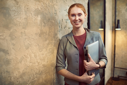Portrait of young office worker holding laptop computer in her hands and smiling at camera in modern office