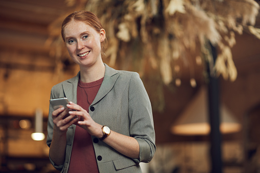 Portrait of young red haired businesswoman standing and smiling at camera while using her mobile phone in cafe