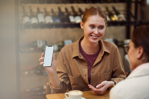 Young red haired woman holding mobile phone and showing something to her friend while they drinking coffee in cafe