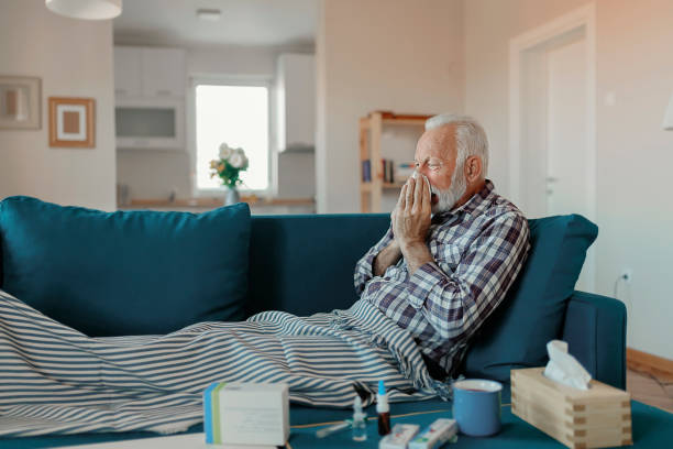 The Flu Cold exhausted senior man with flu wrapped in a warm blanket blowing his nose with a tissue in the livingroom flu virus stock pictures, royalty-free photos & images