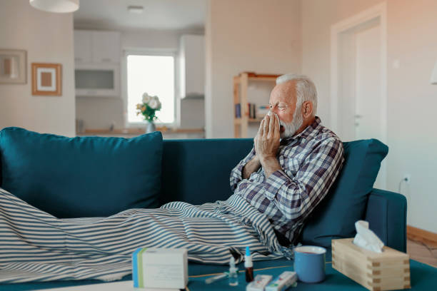 The Flu Cold exhausted senior man with flu wrapped in a warm blanket blowing his nose with a tissue in the livingroom plaid couch stock pictures, royalty-free photos & images