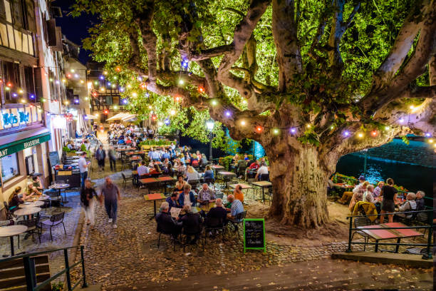 Sidewalk restaurants at night in the Petite France historic quarter in Strasbourg, France. Strasbourg, France - September 14, 2019: People having dinner at night at a sidewalk restaurant on the Quai de la Bruche on the canal in the Petite France quarter, under a 350-year old plane tree. La Petite France stock pictures, royalty-free photos & images