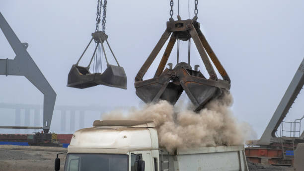 istock Grapple crane unloading cement clinker from ship to truck in river port. Barge delivery of dusty bulk cargo. Gantry crane unloading and loading in Harbor, outdoors. Logistics 1184221214