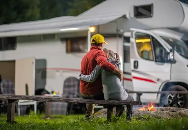RV Campsite Family Time. Father Hugging His Daughter on Wooden Bench in Front of Campfire and Motorhome.
