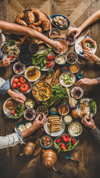 Turkish breakfast table. Flat-lay of Turkish family eating traditional pastries, vegetables, greens, cheeses, fried eggs, jams and tea in copper pot and tulip glasses over wooden background, top view
