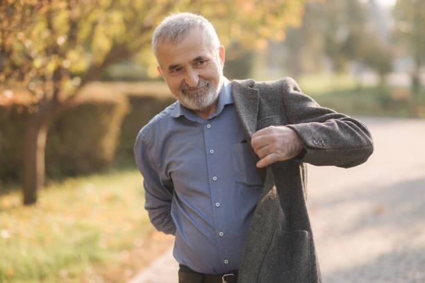 istock Handsome elderly man putting on a gray jacket. Old gray-haired bearded man walk in the autumn park. Yellow background 1184252947