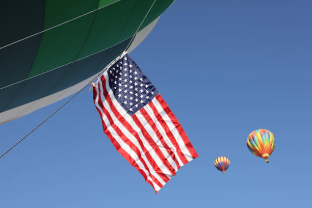 An American flag hangs from a hot air balloon mid-frame in a horizontal composition.  Two other colorful balloons ascend in the background against a clear blue sky.