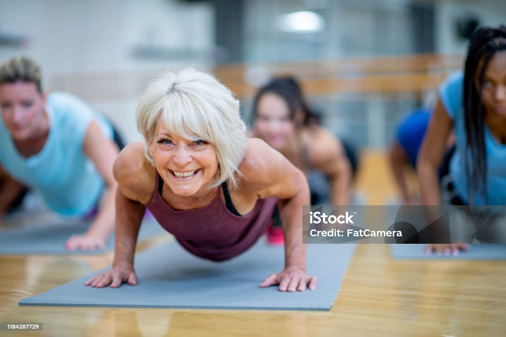 Senior Woman in Fitness Class in a Plank Pose Smiling stock photo An older Caucasian woman is seen holding a plank pose while participating in a  co-ed, multi-ethnic, fitness class.  She is smiling and appearing to enjoy the class. Exercising Stock Photo