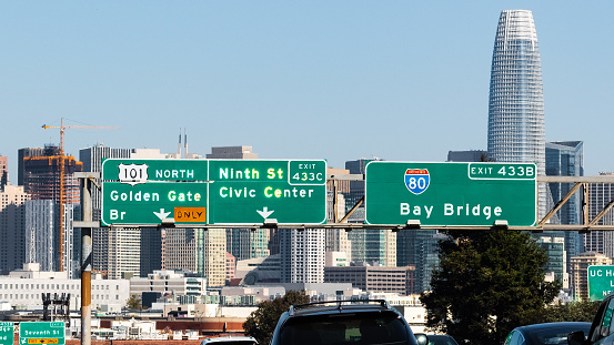 Freeway signage providing direction and information about the upcoming junction; San Francisco downtown skyline visible in the background