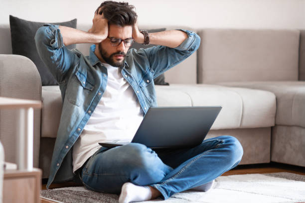 istock Annoyed man holding head and looking at laptop while sitting on floor. 1184324852
