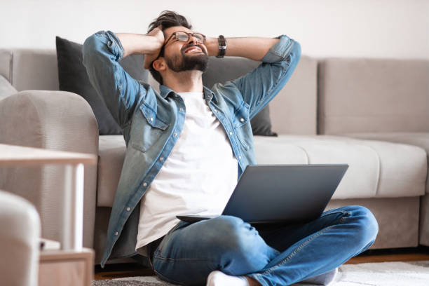istock Annoyed man holding head and looking at laptop while sitting on floor. 1184324900
