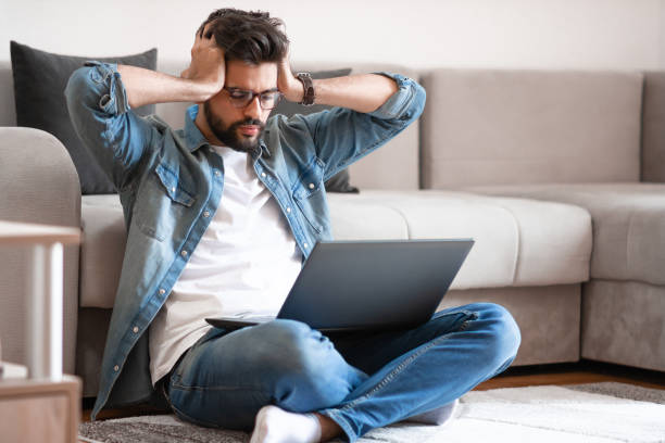 istock Annoyed man holding head and looking at laptop while sitting on floor. 1184324914
