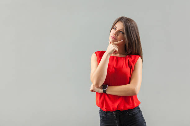 thinking girl. portrait of thoughtful beautiful brunette young woman in red shirt standing, touching her chin, looking away and thinking. - pensive question mark teenager adversity imagens e fotografias de stock